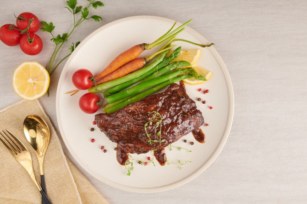 pork roasted grilled spare ribs from a summer bbq served with vegetables asparagus baby carrots fresh tomatoes and spices smoked ribs in white plate on stone surface top view