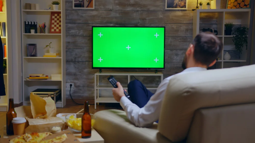 Qual é a melhor tv por assinatura do Brasil hoje 2 back view young man sitting couch front tv with green screen using remote control